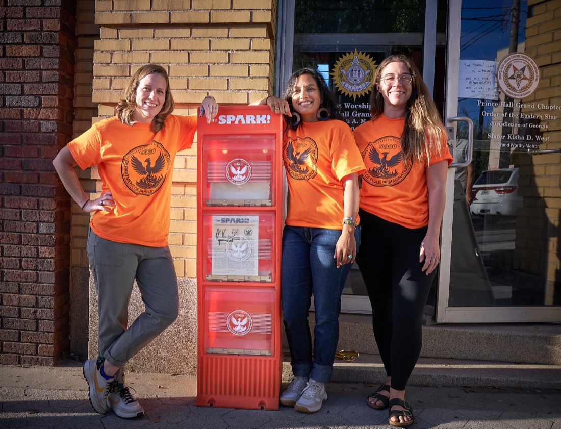 Three DPRI staff members posing beside a fully stocked Spark newsstand